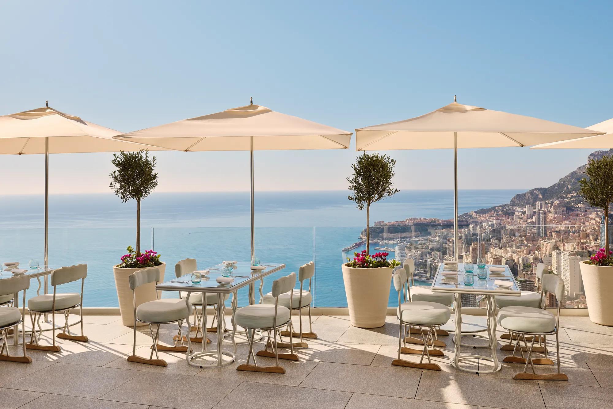 Terrace restaurant with white parasols, potted olive trees, and neatly set tables, overlooking the sea and city coastline under a clear blue sky.