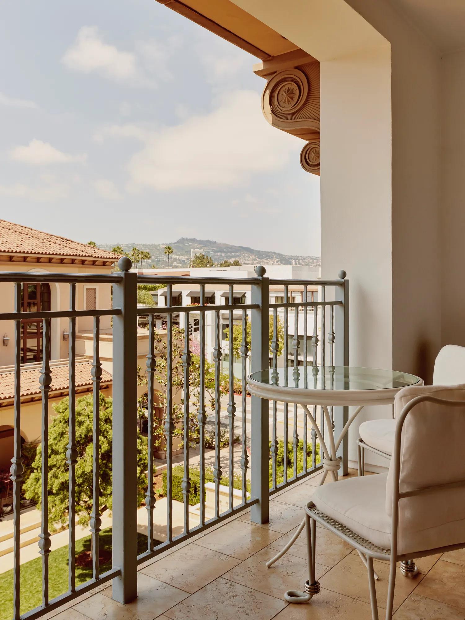 Private balcony with round glass-top table and two cushioned chairs, metal balustrade, and courtyard and hillside views.