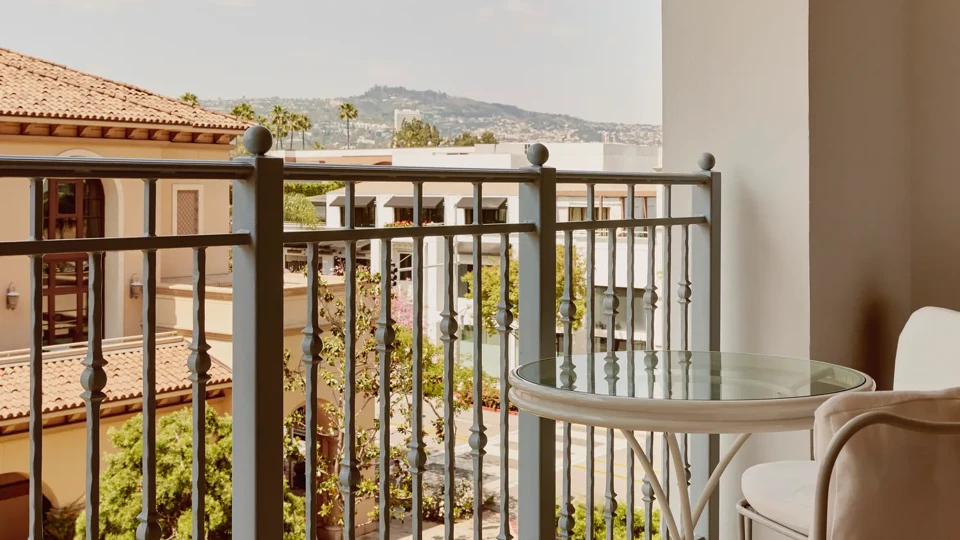 Private balcony with round glass-top table and two cushioned chairs, metal balustrade, and courtyard and hillside views.