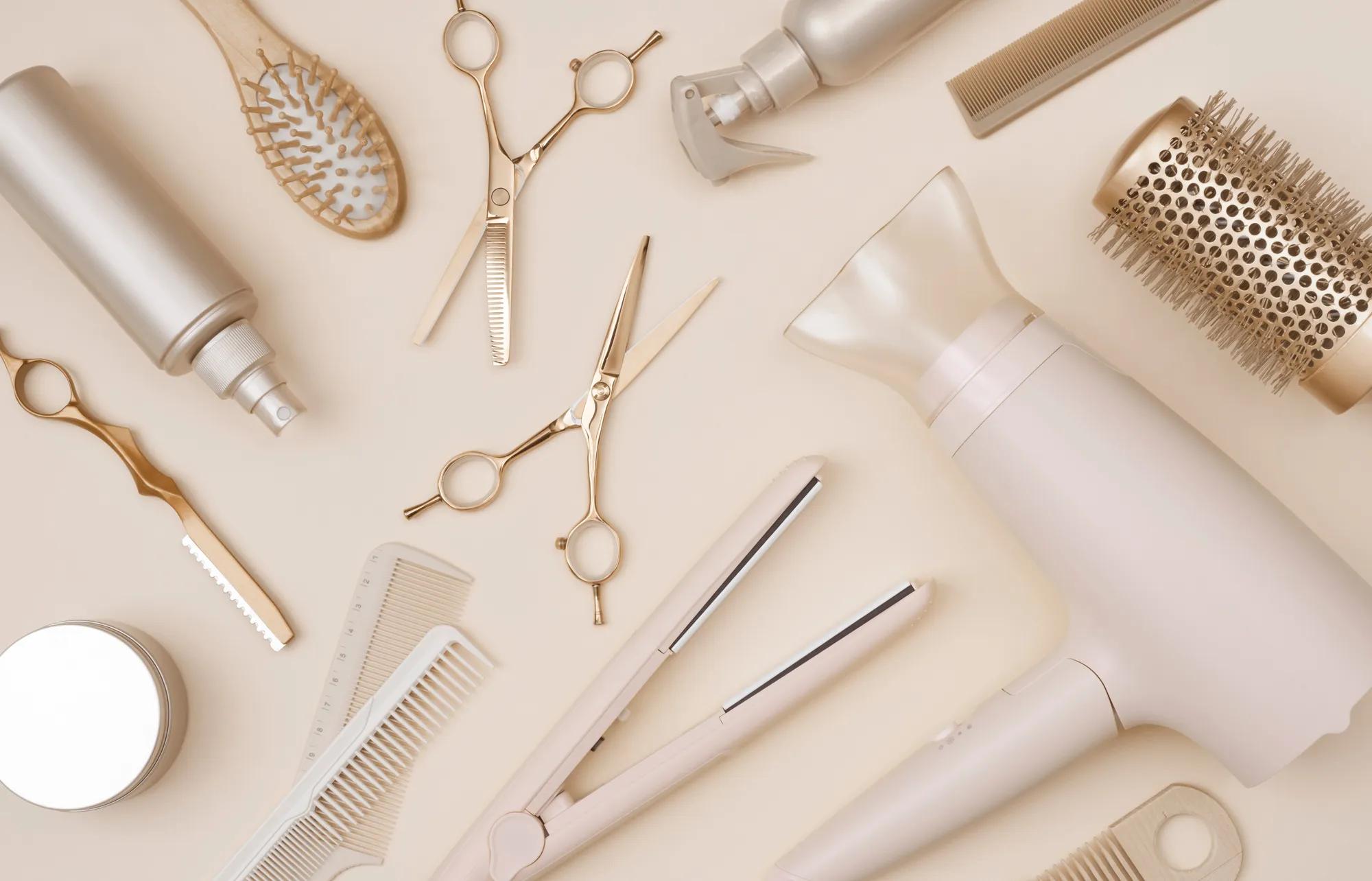 Luxury hair styling tools including a blow dryer, flat iron, salon shears, brushes, combs, and styling products arranged on a soft neutral background.