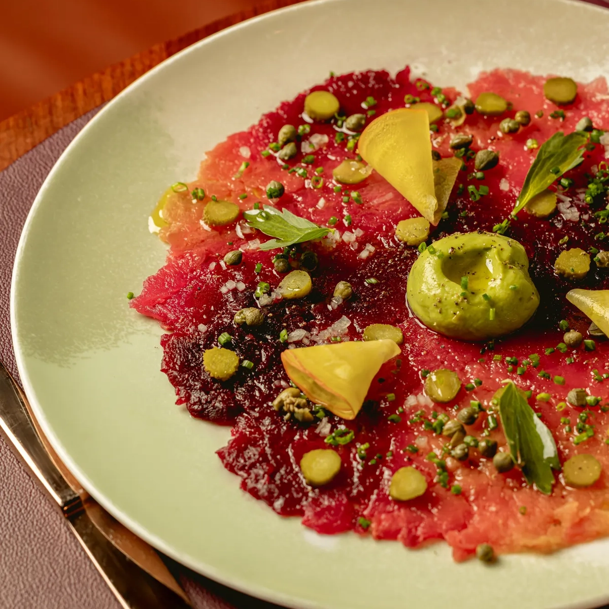 Heirloom beet carpaccio topped with avocado purée, herbs and seeds, served on a pale green plate.