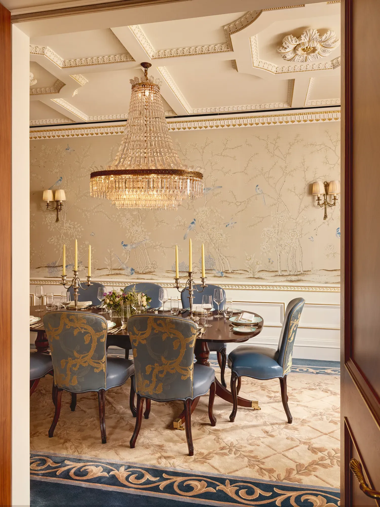 Formal dining room with a polished oval table set for eight, blue and gold upholstered chairs, a grand crystal chandelier, and hand-painted wallpaper depicting trees and birds in soft tones.