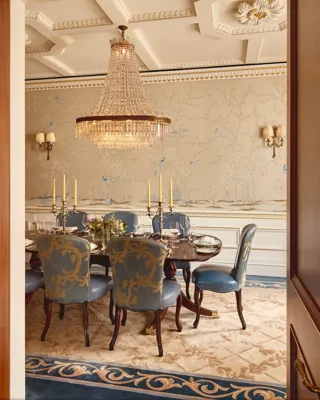 Formal dining room with a polished oval table set for eight, blue and gold upholstered chairs, a grand crystal chandelier, and hand-painted wallpaper depicting trees and birds in soft tones.