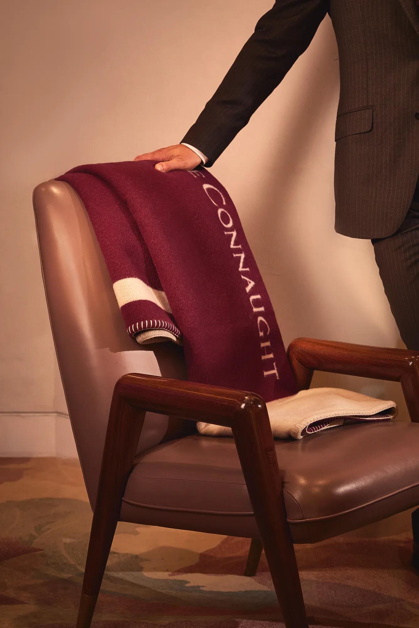 A suited hand rests on a leather chair draped with a folded burgundy blanket embroidered with “Connaught” in white.