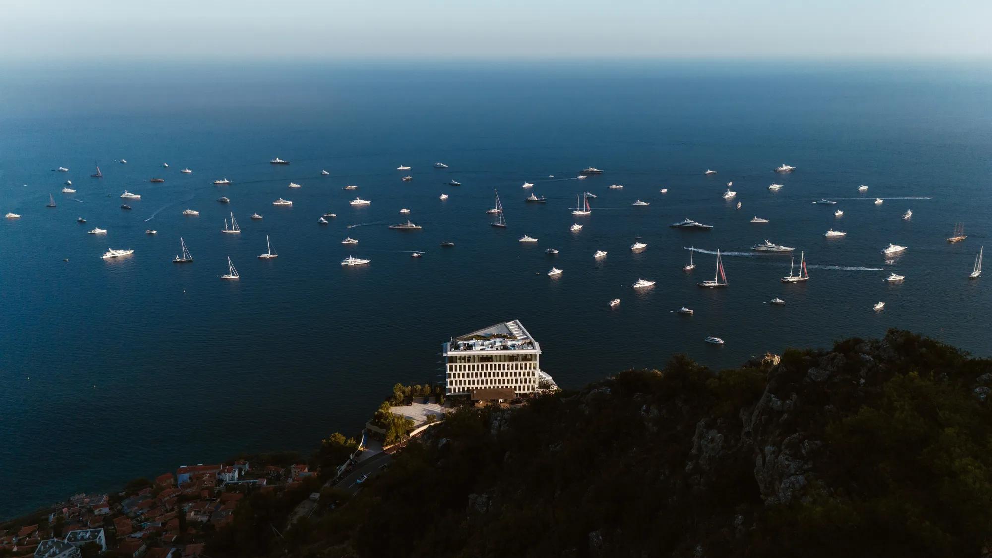Aerial view of seaside hotel on cliff with numerous boats scattered across deep blue Mediterranean waters
