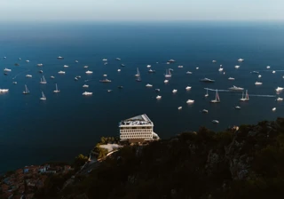 Aerial view of seaside hotel on cliff with numerous boats scattered across deep blue Mediterranean waters