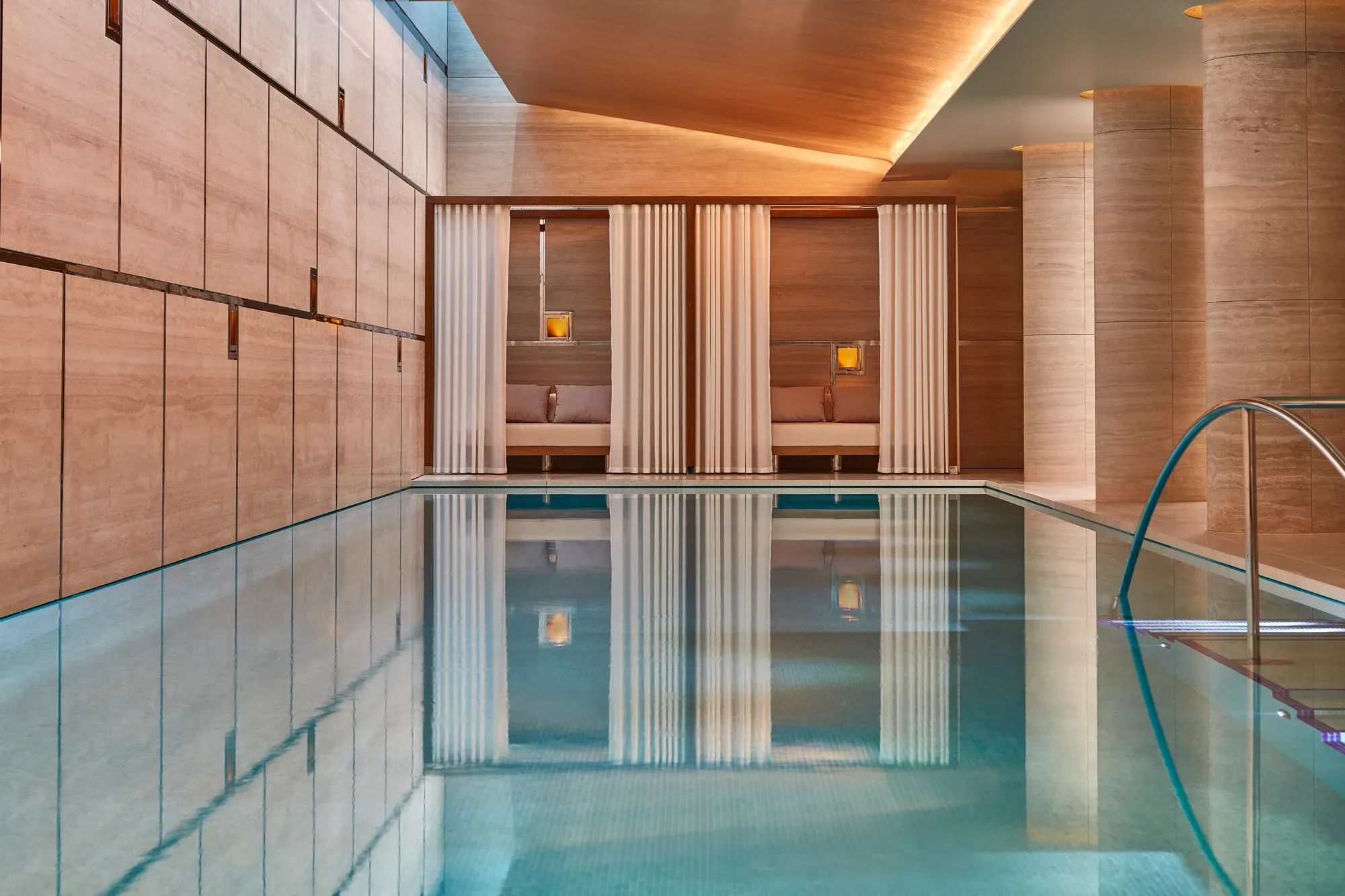 Indoor pool with calm turquoise water, stone walls, and tall columns, featuring curtained cabana seating areas softly lit with warm ambient lighting.