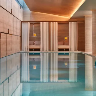 Indoor pool with calm turquoise water, stone walls, and tall columns, featuring curtained cabana seating areas softly lit with warm ambient lighting.