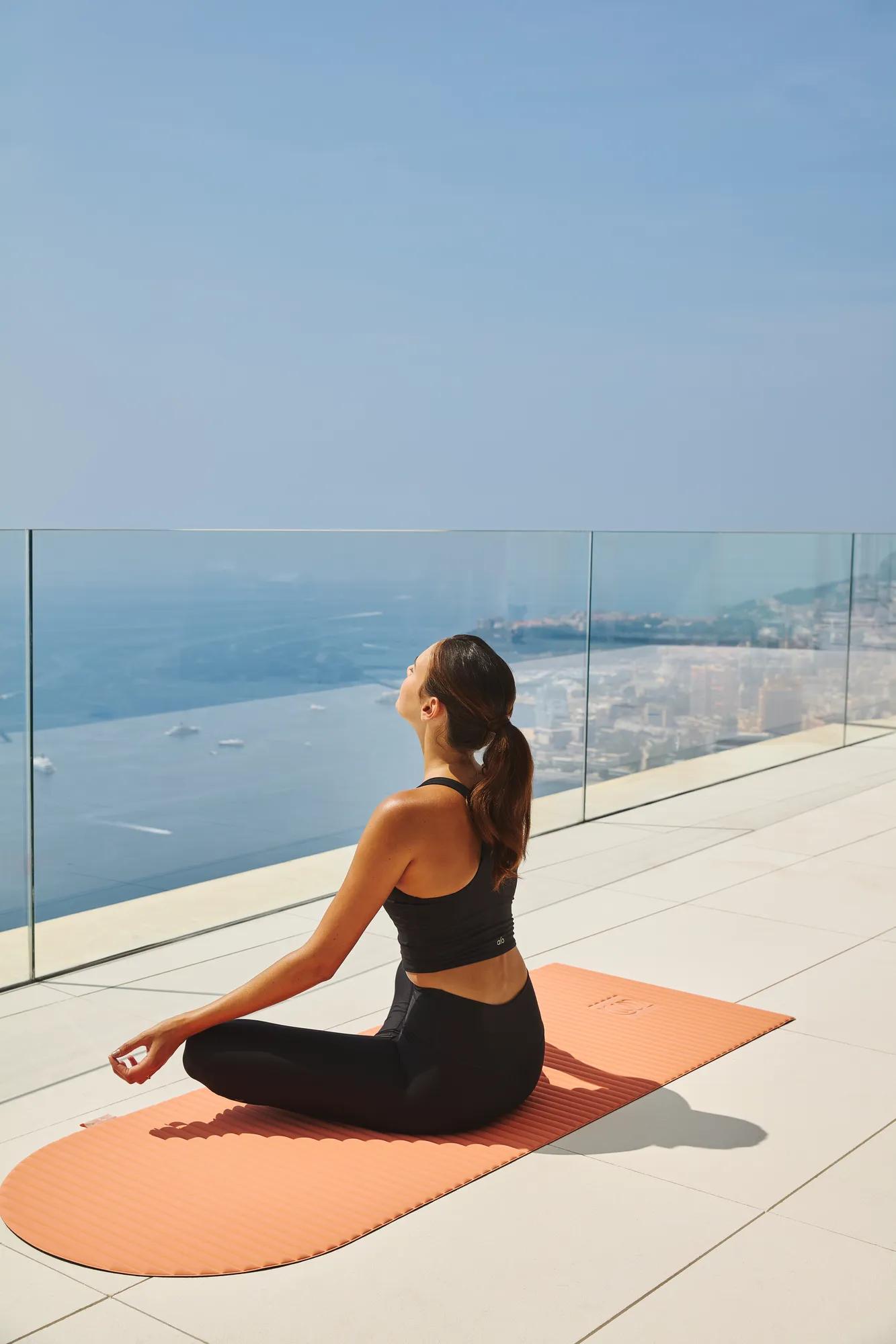 Woman practising yoga on an orange mat, seated cross-legged on a terrace with glass railing, overlooking sea and coastal city views.
