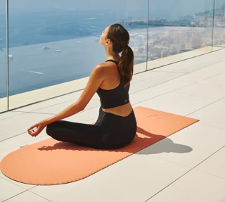 Woman practising yoga on an orange mat, seated cross-legged on a terrace with glass railing, overlooking sea and coastal city views.