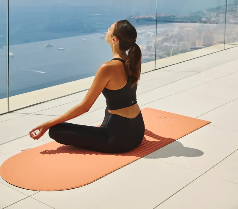 Woman practising yoga on an orange mat, seated cross-legged on a terrace with glass railing, overlooking sea and coastal city views.