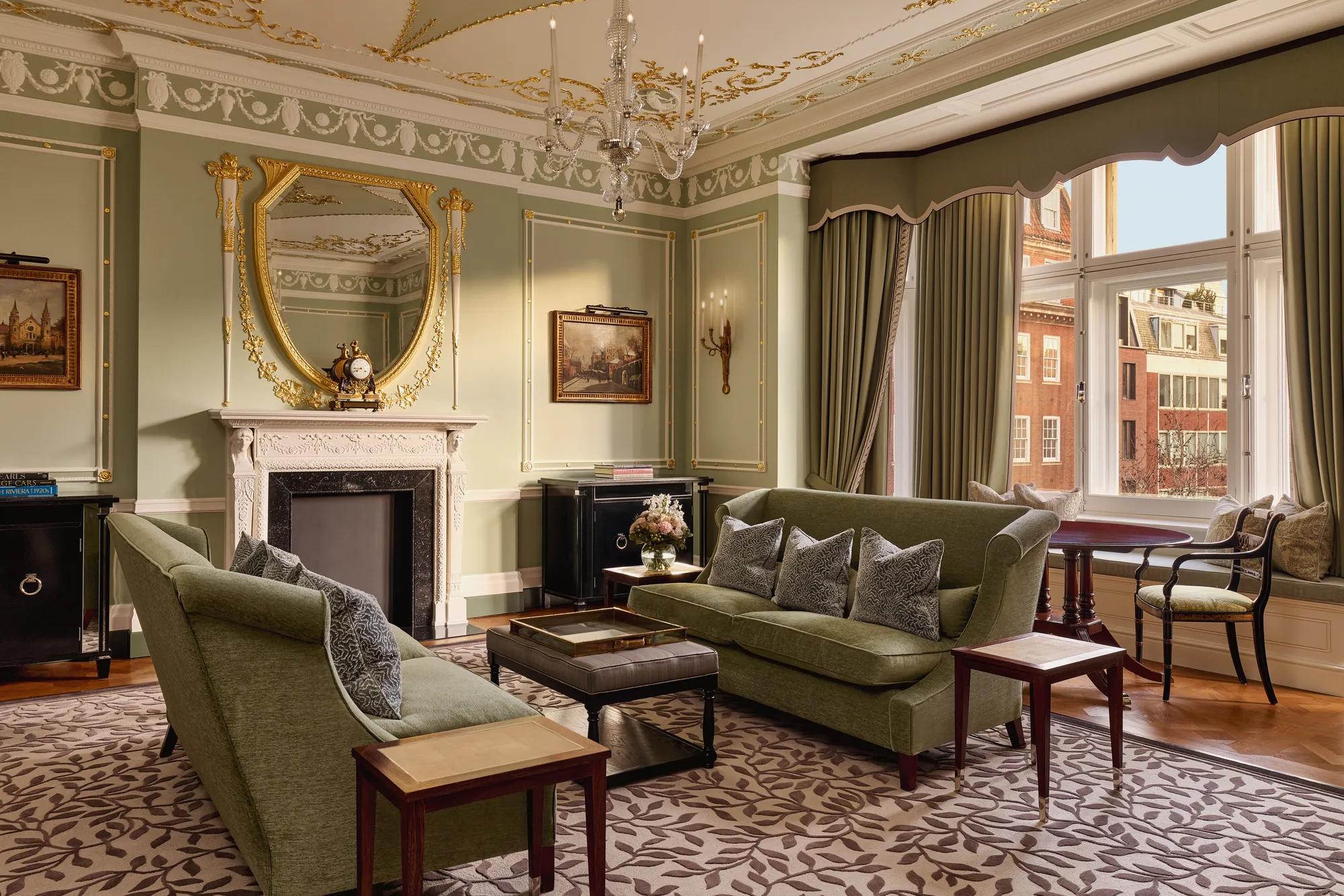 Elegant sitting room with green sofas, ornate fireplace, gold-framed mirror, and large windows with city views.