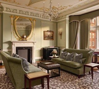 Elegant sitting room with green sofas, ornate fireplace, gold-framed mirror, and large windows with city views.