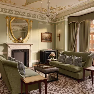 Elegant sitting room with green sofas, ornate fireplace, gold-framed mirror, and large windows with city views.