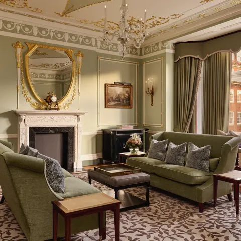 Elegant sitting room with green sofas, ornate fireplace, gold-framed mirror, and large windows with city views.