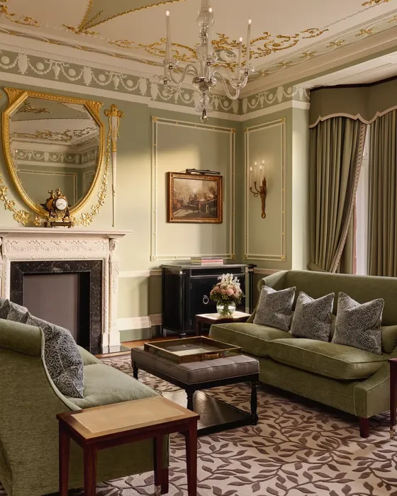 Elegant sitting room with green sofas, ornate fireplace, gold-framed mirror, and large windows with city views.