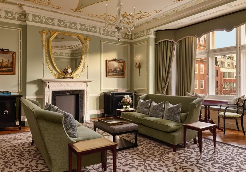 Elegant sitting room with green sofas, ornate fireplace, gold-framed mirror, and large windows with city views.