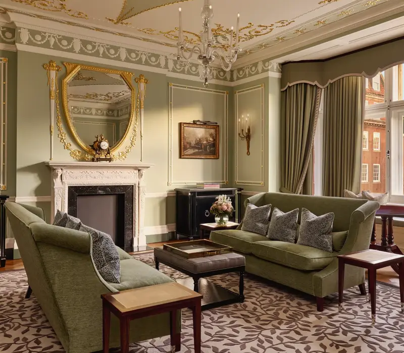 Elegant sitting room with green sofas, ornate fireplace, gold-framed mirror, and large windows with city views.