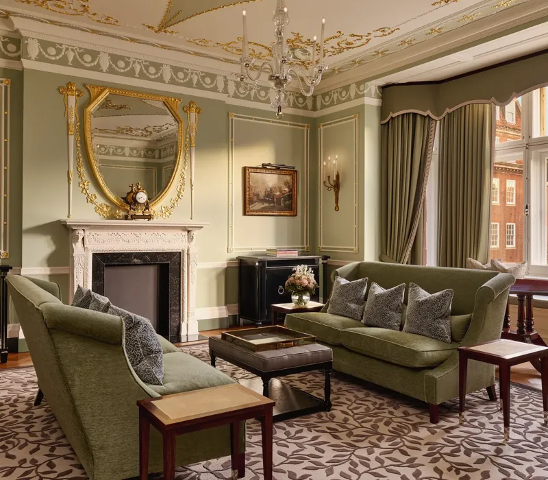 Elegant sitting room with green sofas, ornate fireplace, gold-framed mirror, and large windows with city views.