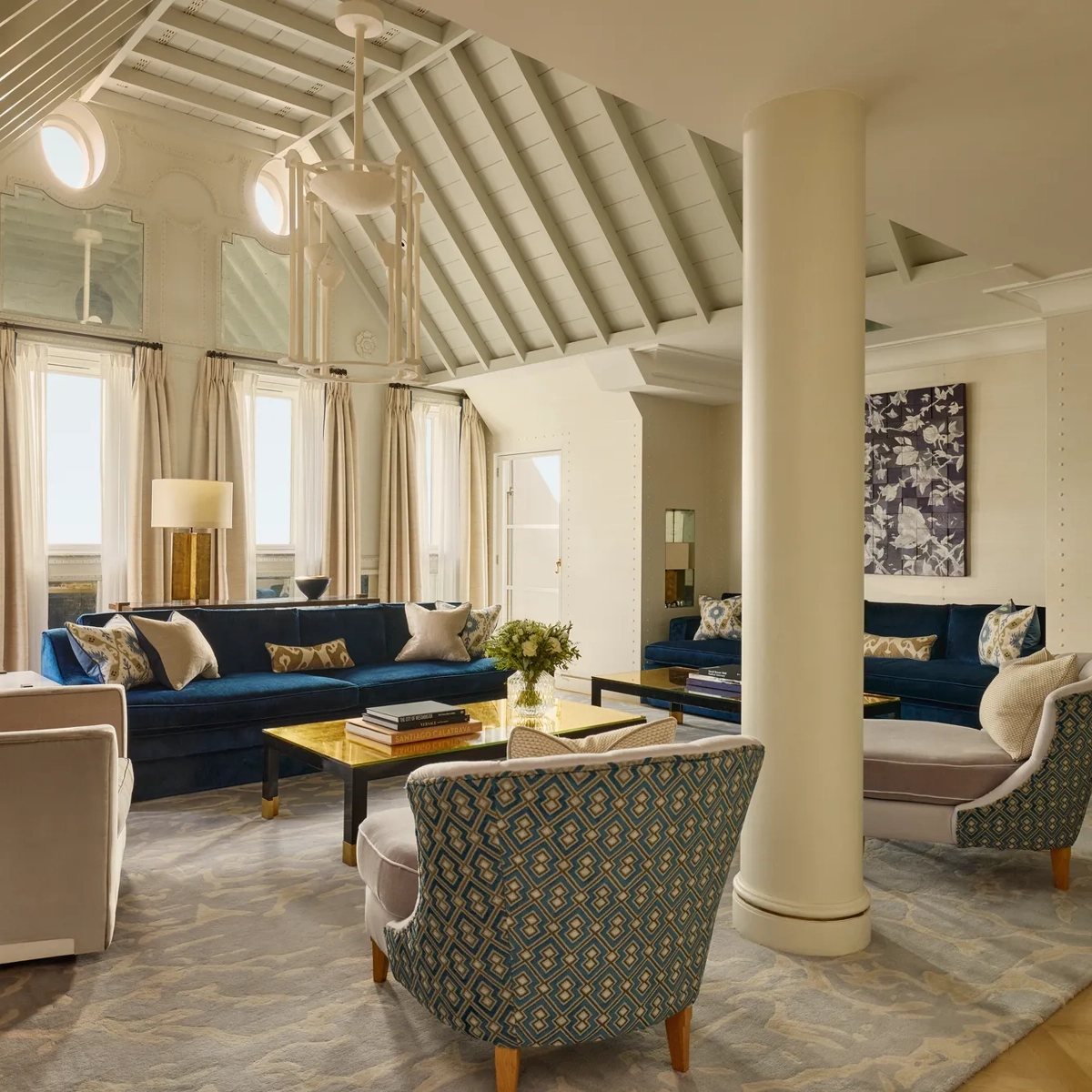 A bright living room with vaulted ceilings, blue velvet sofas, patterned armchairs, and soft neutral curtains, featuring contemporary décor, natural light, and a mix of classic and modern design details.