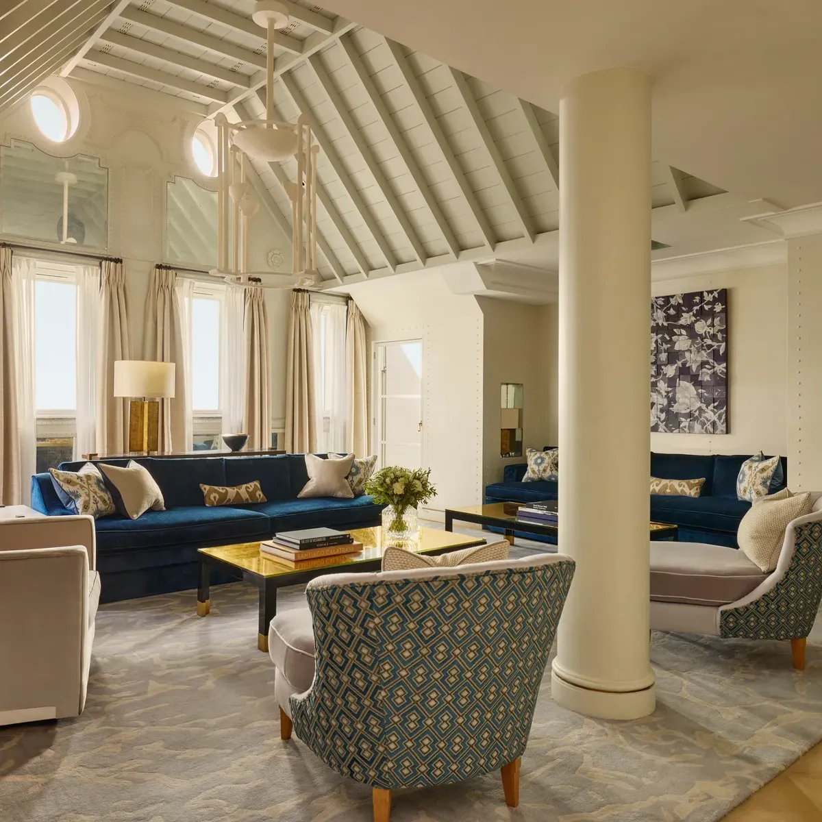 A bright living room with vaulted ceilings, blue velvet sofas, patterned armchairs, and soft neutral curtains, featuring contemporary décor, natural light, and a mix of classic and modern design details.