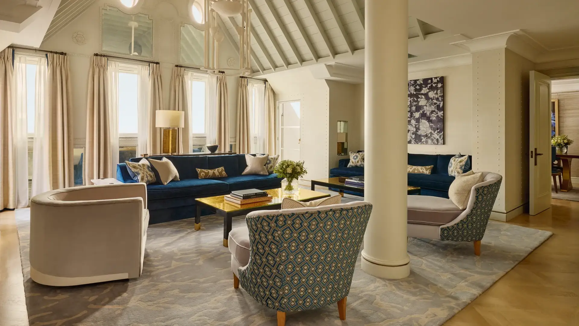 A bright living room with vaulted ceilings, blue velvet sofas, patterned armchairs, and soft neutral curtains, featuring contemporary décor, natural light, and a mix of classic and modern design details.