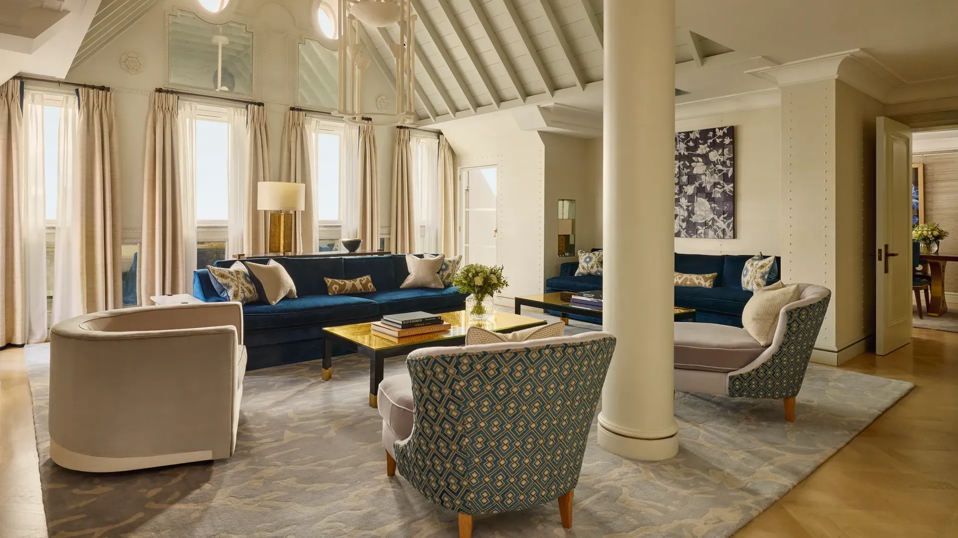 A bright living room with vaulted ceilings, blue velvet sofas, patterned armchairs, and soft neutral curtains, featuring contemporary décor, natural light, and a mix of classic and modern design details.
