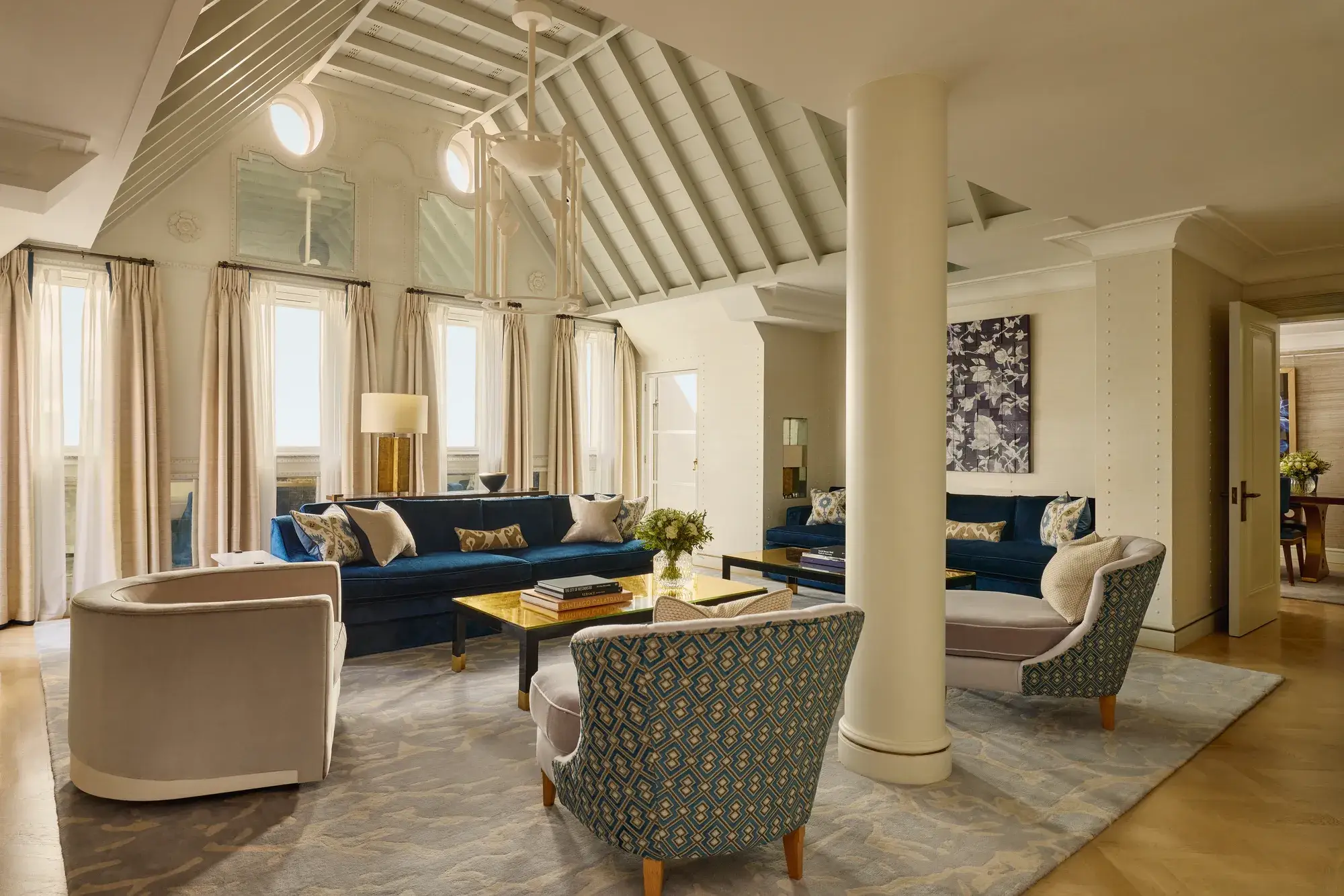 A bright living room with vaulted ceilings, blue velvet sofas, patterned armchairs, and soft neutral curtains, featuring contemporary décor, natural light, and a mix of classic and modern design details.