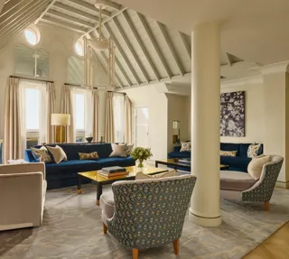 A bright living room with vaulted ceilings, blue velvet sofas, patterned armchairs, and soft neutral curtains, featuring contemporary décor, natural light, and a mix of classic and modern design details.