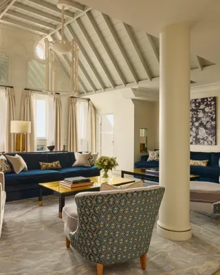A bright living room with vaulted ceilings, blue velvet sofas, patterned armchairs, and soft neutral curtains, featuring contemporary décor, natural light, and a mix of classic and modern design details.