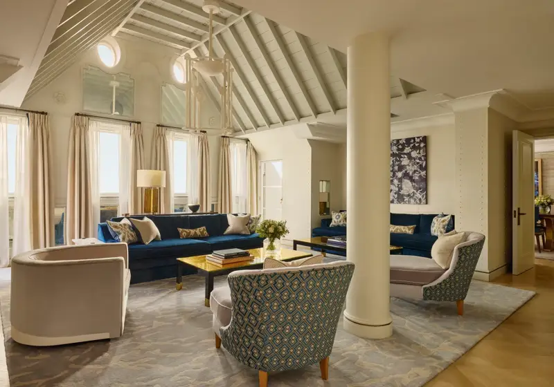 A bright living room with vaulted ceilings, blue velvet sofas, patterned armchairs, and soft neutral curtains, featuring contemporary décor, natural light, and a mix of classic and modern design details.