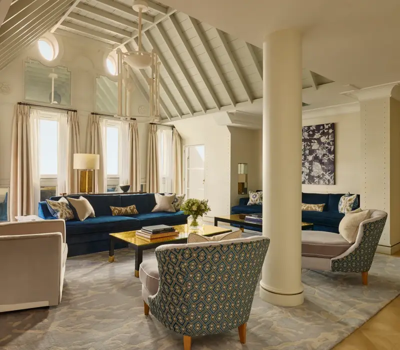 A bright living room with vaulted ceilings, blue velvet sofas, patterned armchairs, and soft neutral curtains, featuring contemporary décor, natural light, and a mix of classic and modern design details.