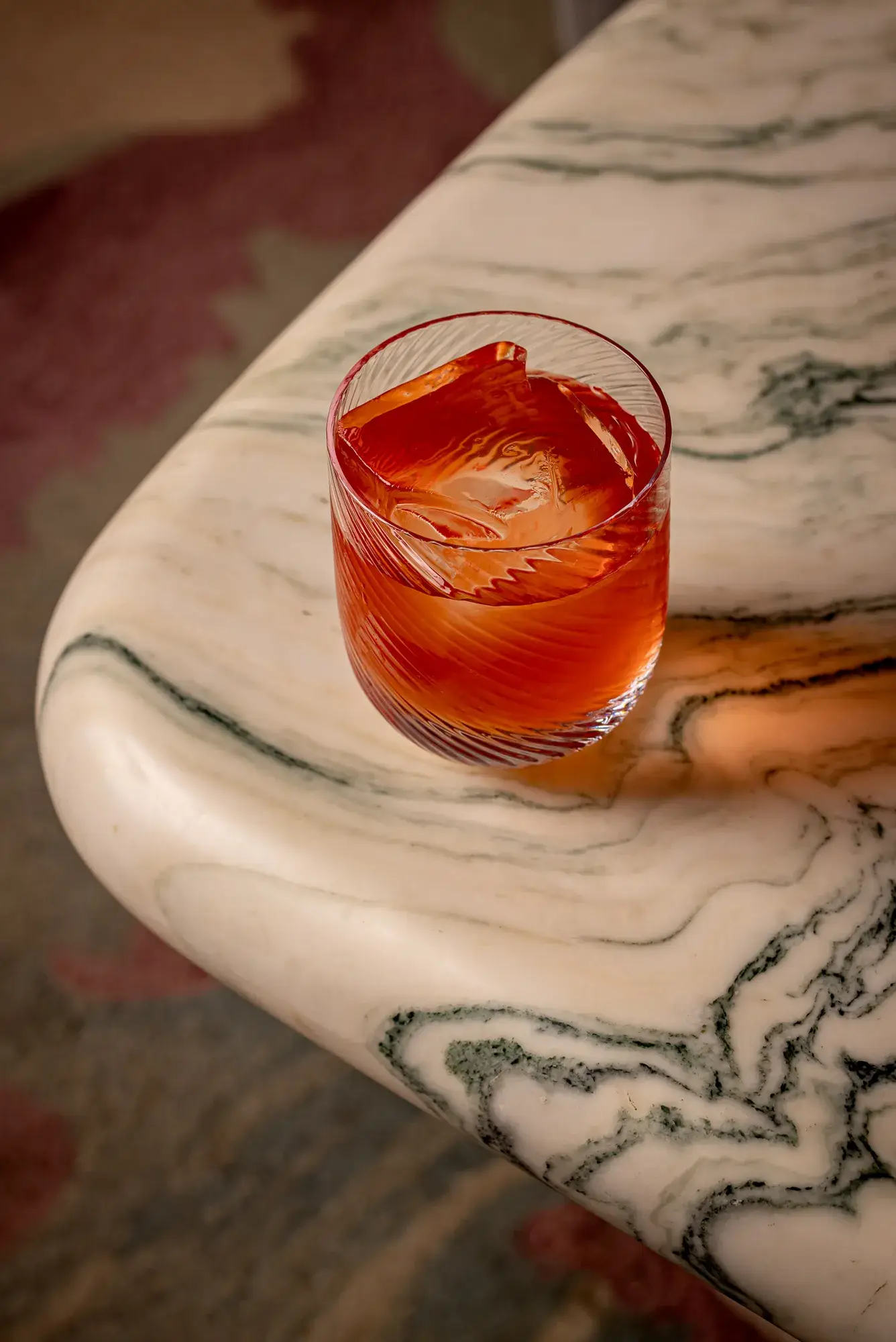 Negroni-style orange cocktail in a patterned glass resting on a marbled table edge.