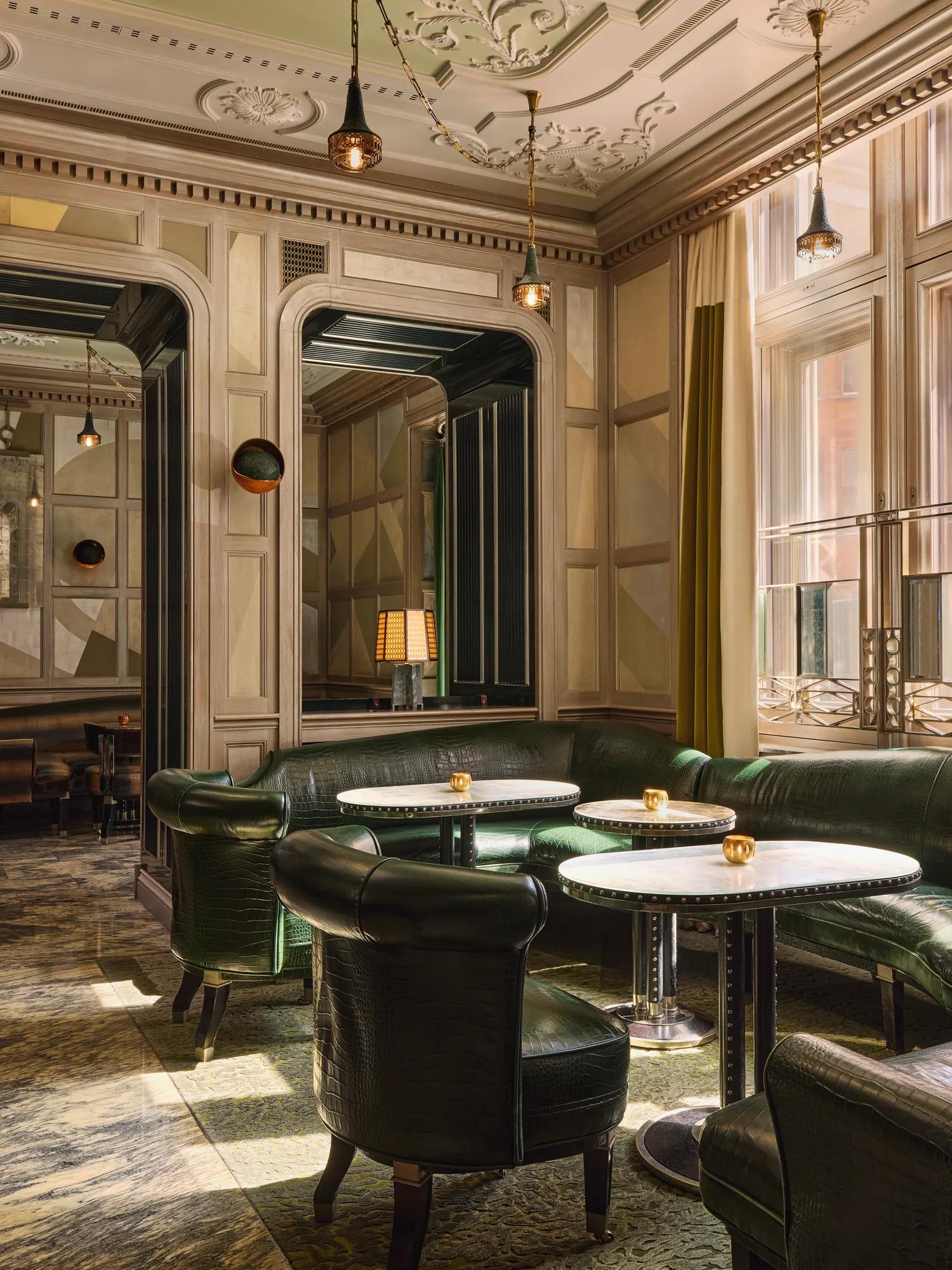 Intimate lounge with green leather seating, round marble tables, and warm light from tall windows.