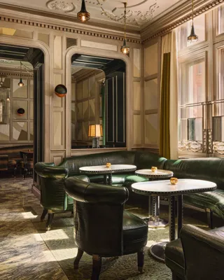 Intimate lounge with green leather seating, round marble tables, and warm light from tall windows.