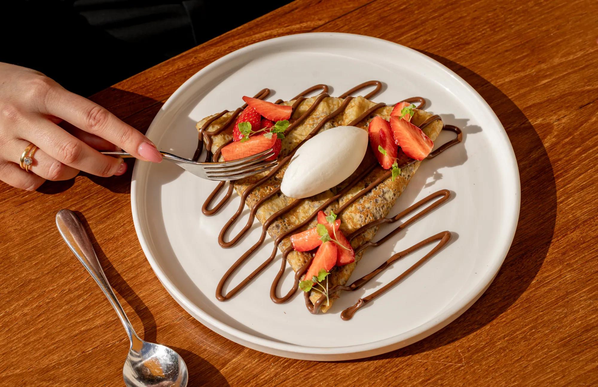 **Alt text (WCAG 2.2 AA):**

A folded crêpe drizzled with chocolate, topped with fresh strawberries and a scoop of vanilla ice cream on a white plate, with a hand reaching for a bite.
