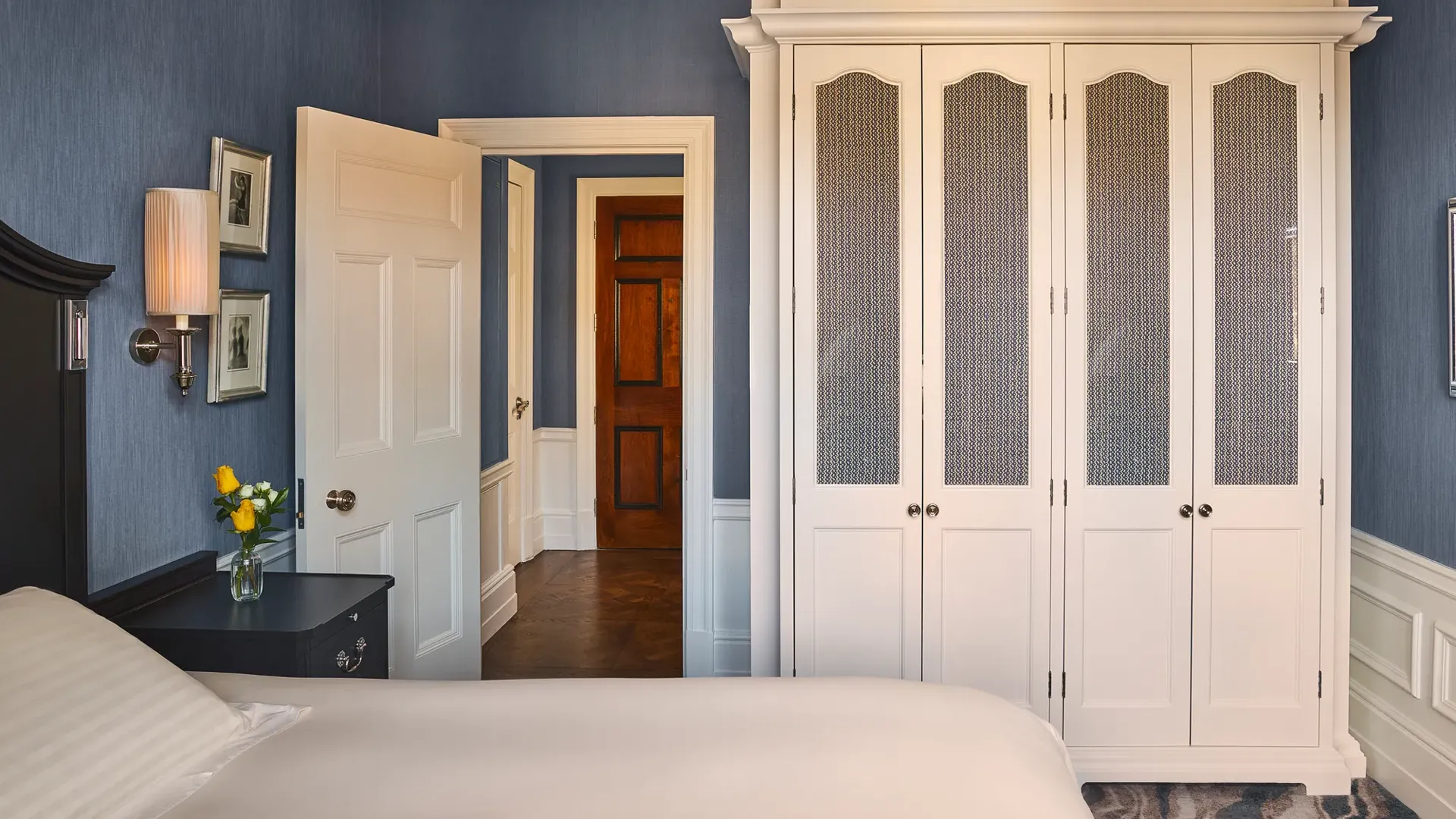 Bedroom with blue walls, white wardrobe, bedside table with lamp and flowers, and open door to hallway.