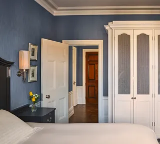 Bedroom with blue walls, white wardrobe, bedside table with lamp and flowers, and open door to hallway.