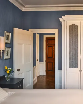 Bedroom with blue walls, white wardrobe, bedside table with lamp and flowers, and open door to hallway.