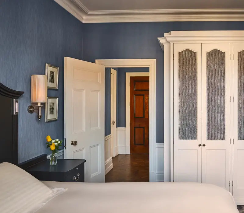Bedroom with blue walls, white wardrobe, bedside table with lamp and flowers, and open door to hallway.