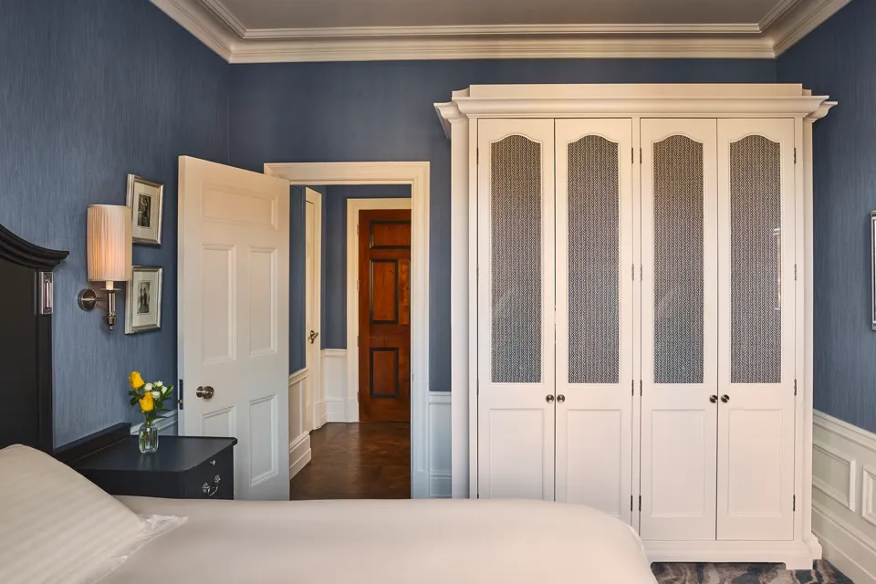 Bedroom with blue walls, white wardrobe, bedside table with lamp and flowers, and open door to hallway.