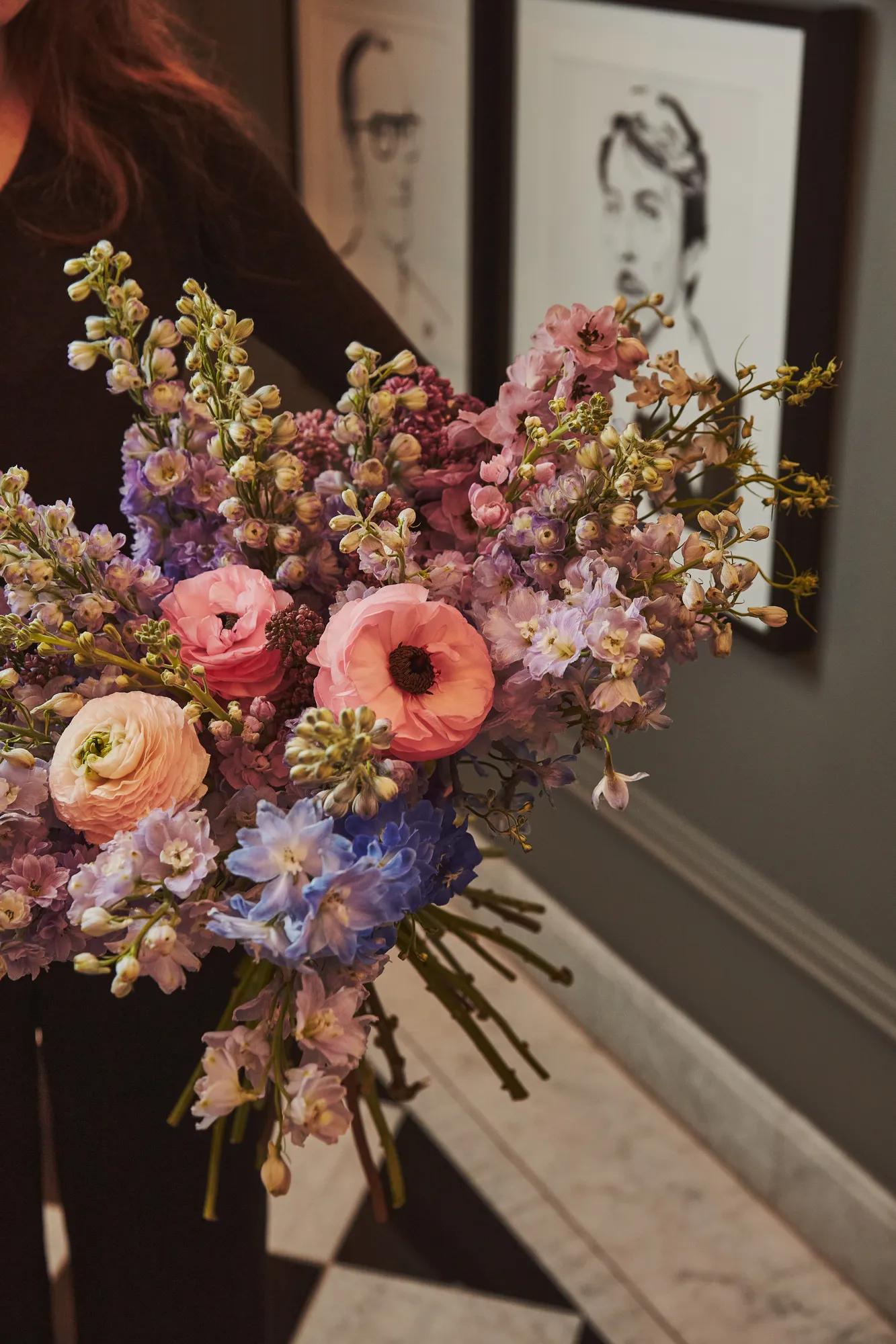 Close-up of a pastel flower bouquet with pink, lilac and blue blooms, held in an elegant hallway with framed portraits.