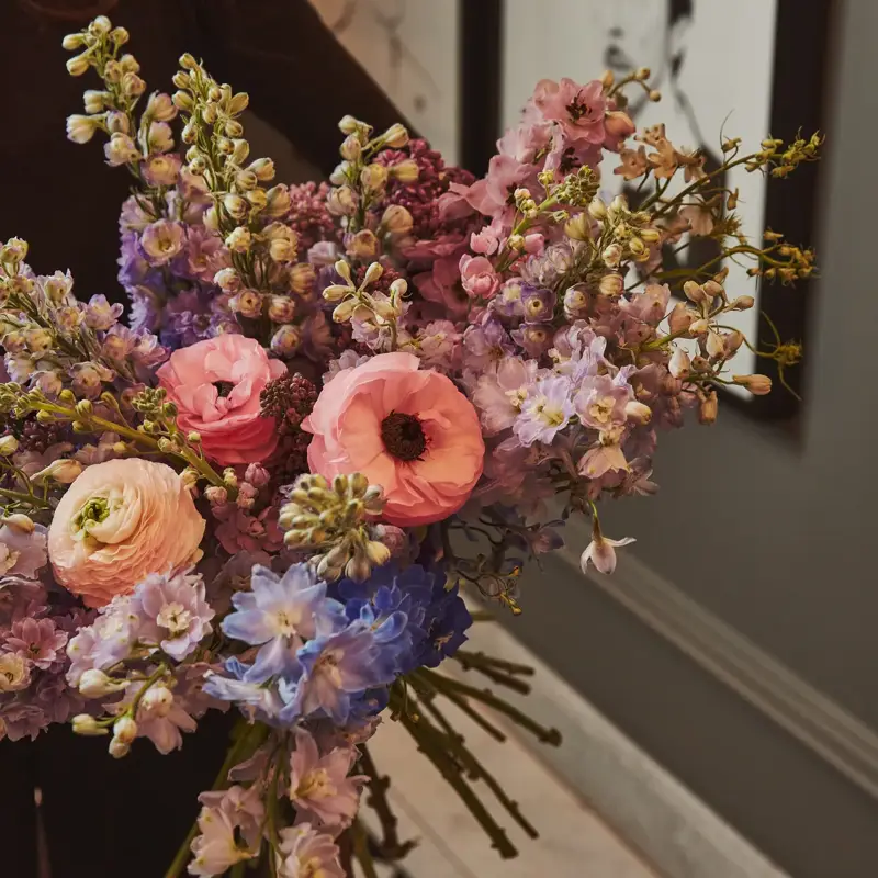 Close-up of a pastel flower bouquet with pink, lilac and blue blooms, held in an elegant hallway with framed portraits.