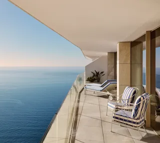 Balcony with lounge chairs and striped armchair overlooking calm sea, featuring glass railing and soft daylight along a modern terrace.