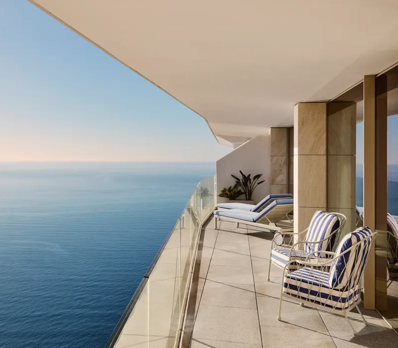 Balcony with lounge chairs and striped armchair overlooking calm sea, featuring glass railing and soft daylight along a modern terrace.