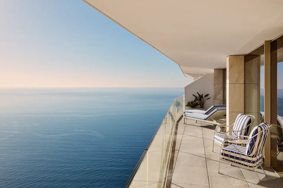 Balcony with lounge chairs and striped armchair overlooking calm sea, featuring glass railing and soft daylight along a modern terrace.