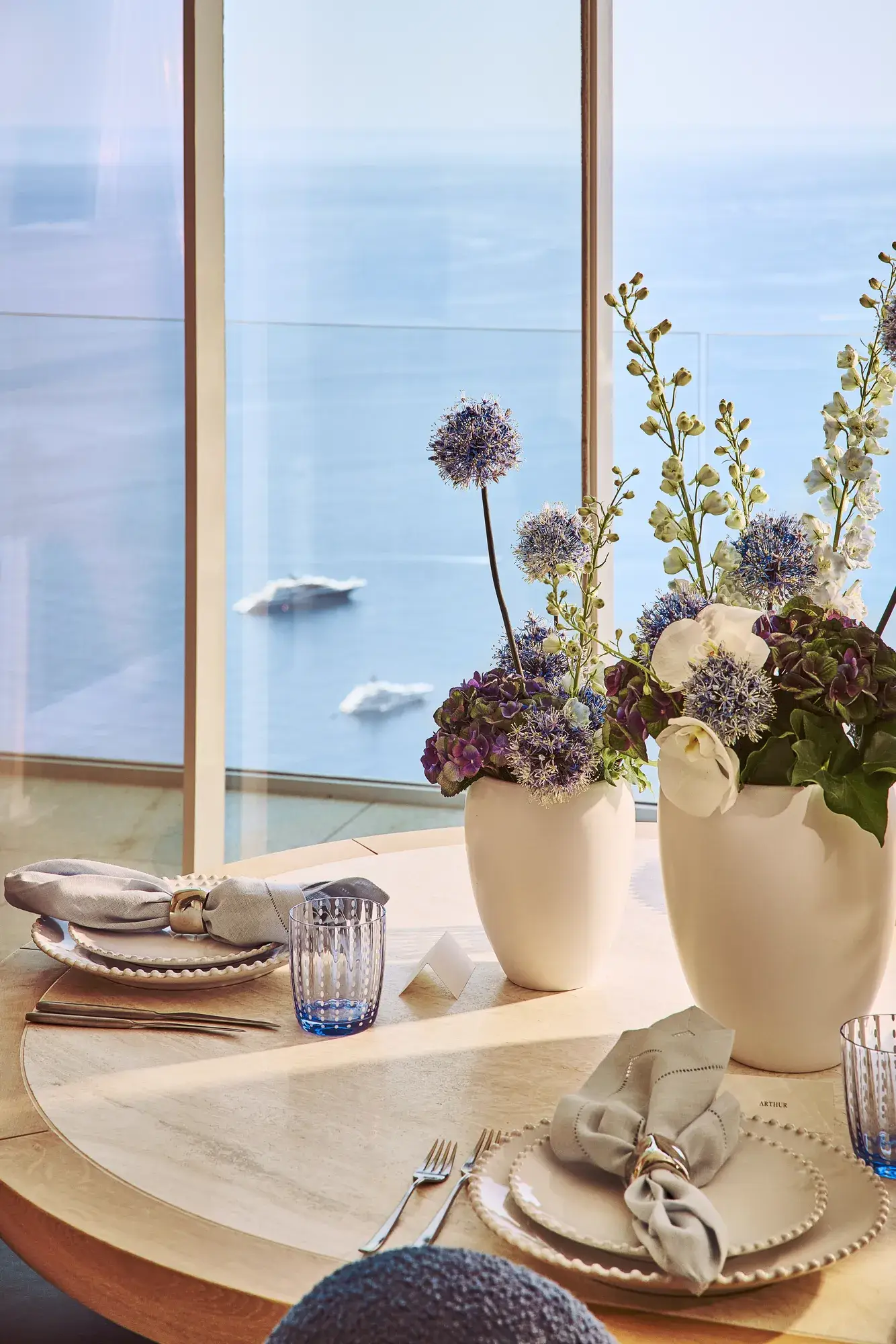 Table ronde élégamment dressée avec des assiettes blanches, des serviettes en lin et des verres bleus, décorée de vases blancs remplis de fleurs violettes et blanches, près d’une fenêtre donnant sur la mer.