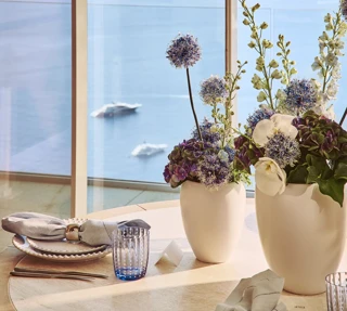 Table ronde élégamment dressée avec des assiettes blanches, des serviettes en lin et des verres bleus, décorée de vases blancs remplis de fleurs violettes et blanches, près d’une fenêtre donnant sur la mer.