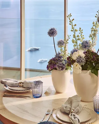 Table ronde élégamment dressée avec des assiettes blanches, des serviettes en lin et des verres bleus, décorée de vases blancs remplis de fleurs violettes et blanches, près d’une fenêtre donnant sur la mer.