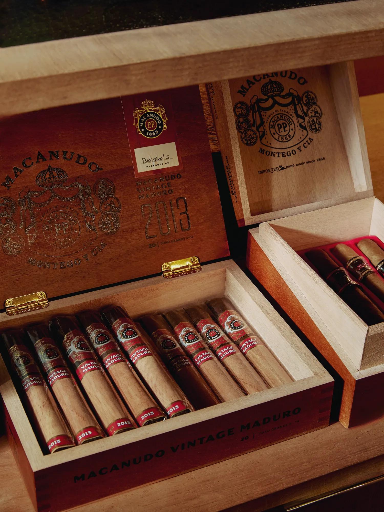 Open Macanudo cigar boxes on a shelf with vintage maduro sticks neatly lined, brass hinges and logos visible.